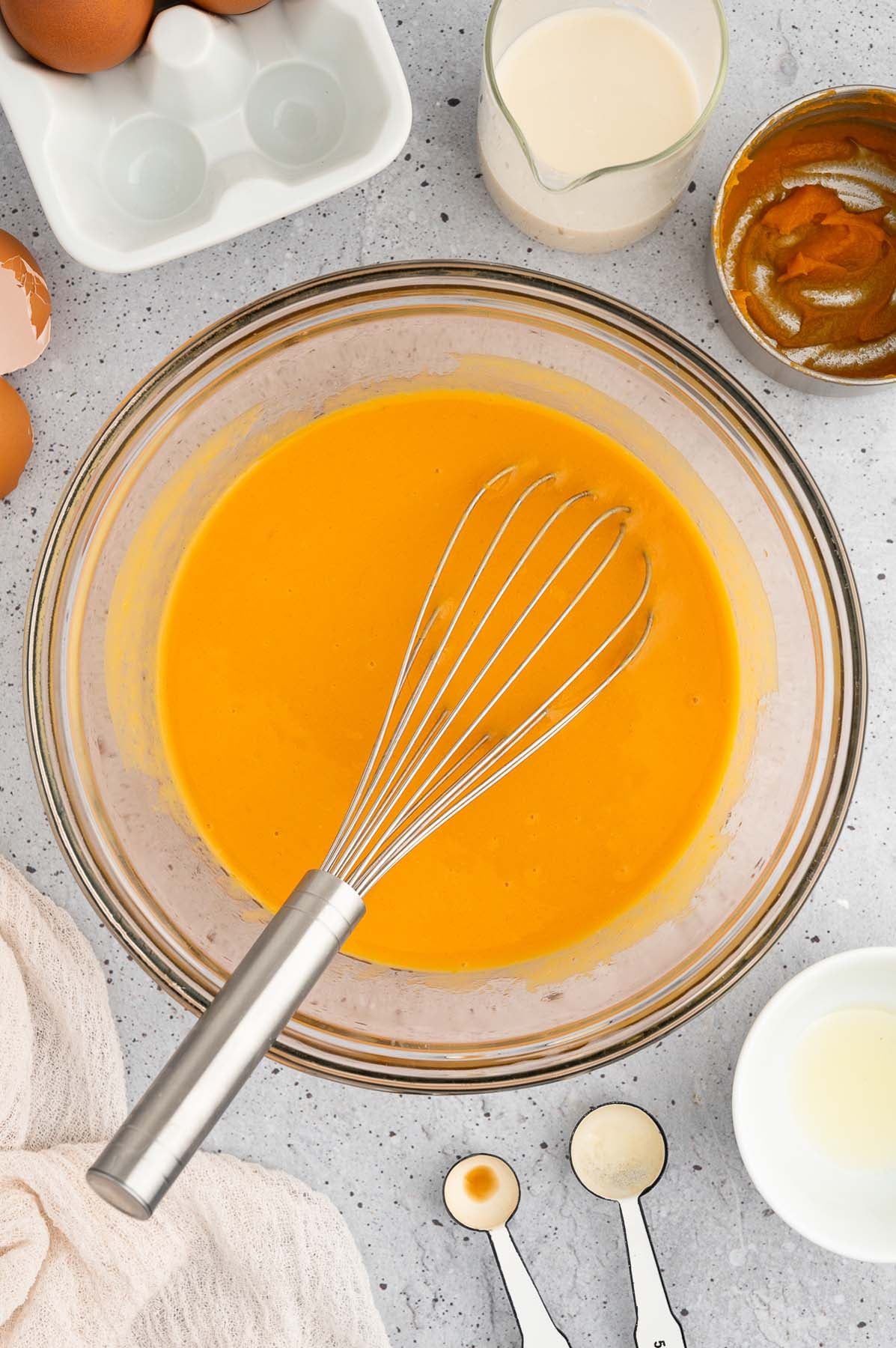 Wet ingredients for sweet potato pancakes in a clear bowl with a whisk on a grey background with ingredients on the side