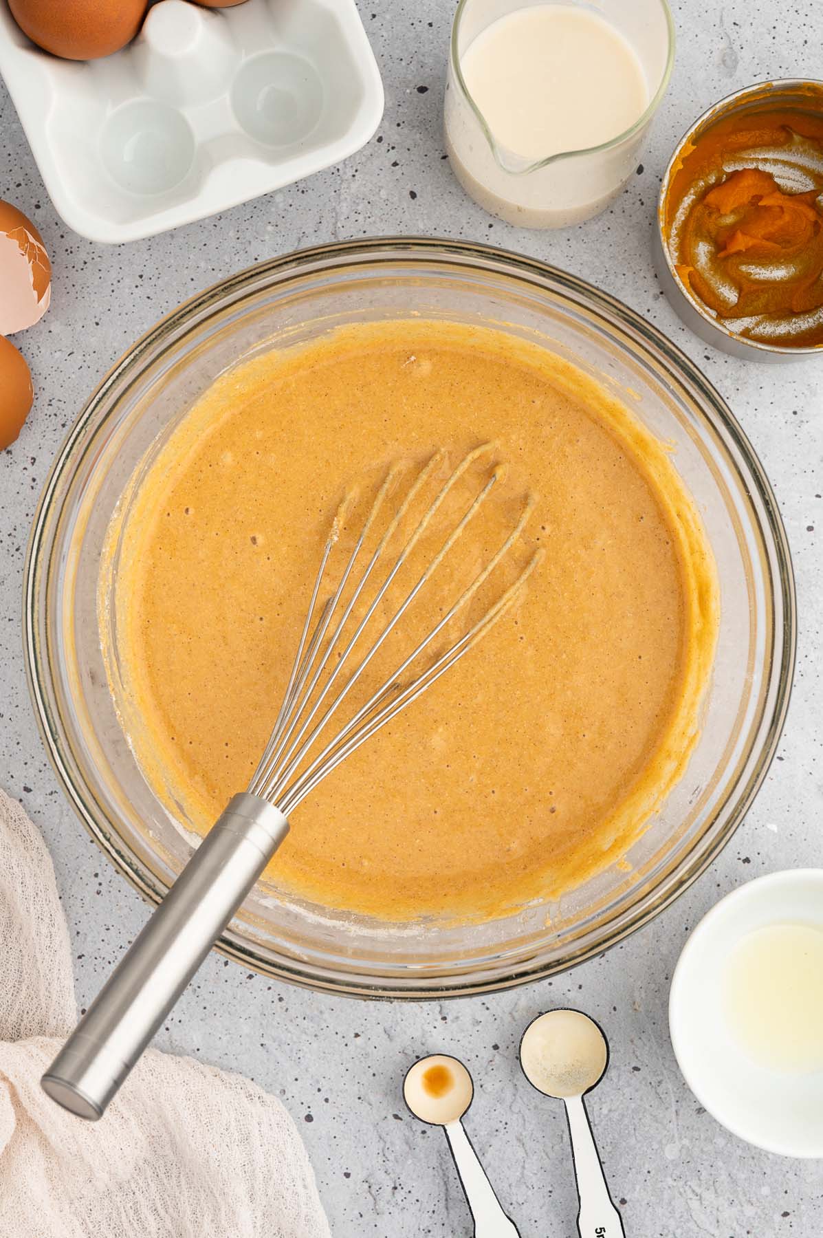 Pancake Batter for sweet potato pancakes in a clear bowl with a whisk in it on a grey background with ingredients on the side