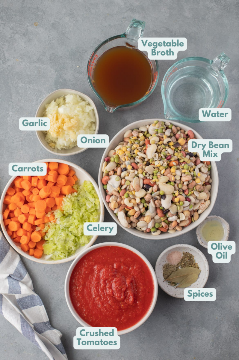 Ingredients for 15 bean soup in white bowls on a grey background: olive oil, onion, celery, carrots, salt, pepper, bay leaf, dried basil, dried oregano, dried parsley, crushed tomatoes, broth (veggie or chicken broth), water, 15 bean soup mix 