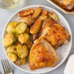 An overhead shot of a plate with baked chicken thighs, roasted fingerling potatoes, and parmesan brussel sprouts with a sheet pan of more chicken, a glass of wine, and gold silverware on the side