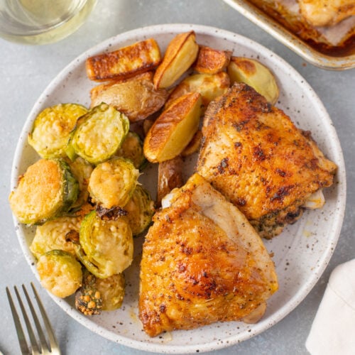 An overhead shot of a plate with baked chicken thighs, roasted fingerling potatoes, and parmesan brussel sprouts with a sheet pan of more chicken, a glass of wine, and gold silverware on the side