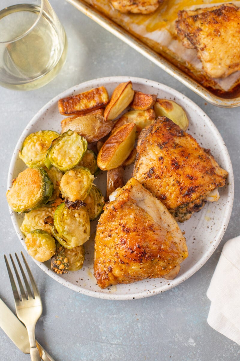 An overhead shot of a plate with baked chicken thighs, roasted fingerling potatoes, and parmesan brussel sprouts with a sheet pan of more chicken, a glass of wine, and gold silverware on the side