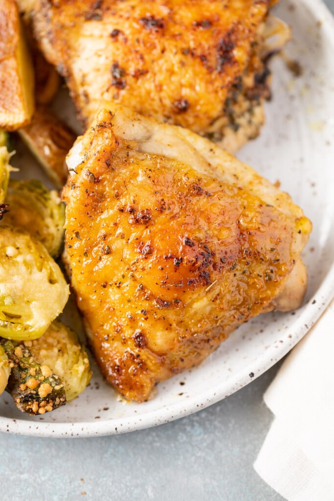 A close up of a baked chicken thigh on a white speckled plate with brussel sprouts on the side