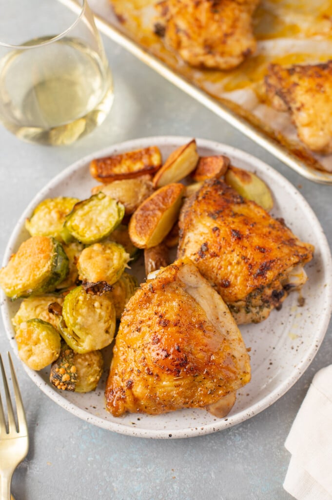 A white speckled plate with baked chicken thighs, roasted fingerling potatoes, and parmesan brussel sprouts with a sheet pan of more chicken, a glass of wine, and a gold fork on the side