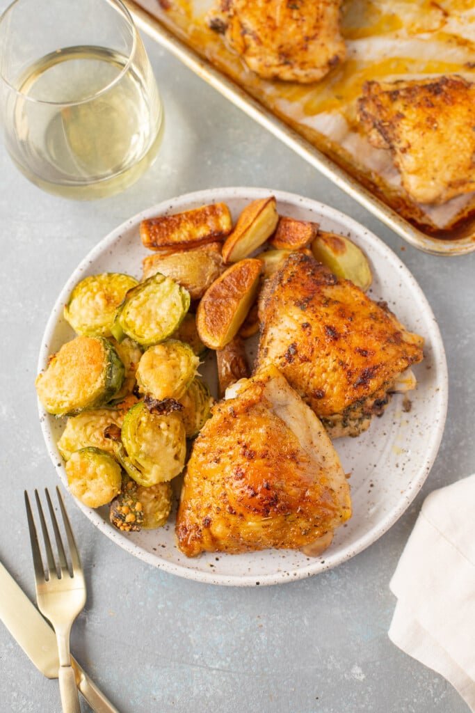 An overhead shot of a white speckled plate with baked chicken thighs, roasted fingerling potatoes, and parmesan brussel sprouts with a sheet pan of more chicken, a glass of wine, and gold silverware on the side