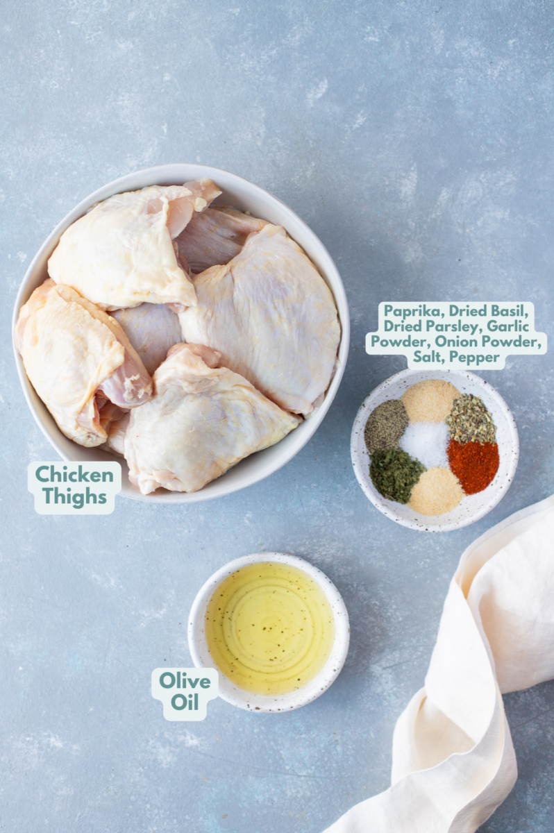 Ingredients for baked chicken thighs in white bowls: bone in chicken thighs, olive oil, paprika, dried basil, dried parsley, garlic powder, onion powder, salt and black pepper