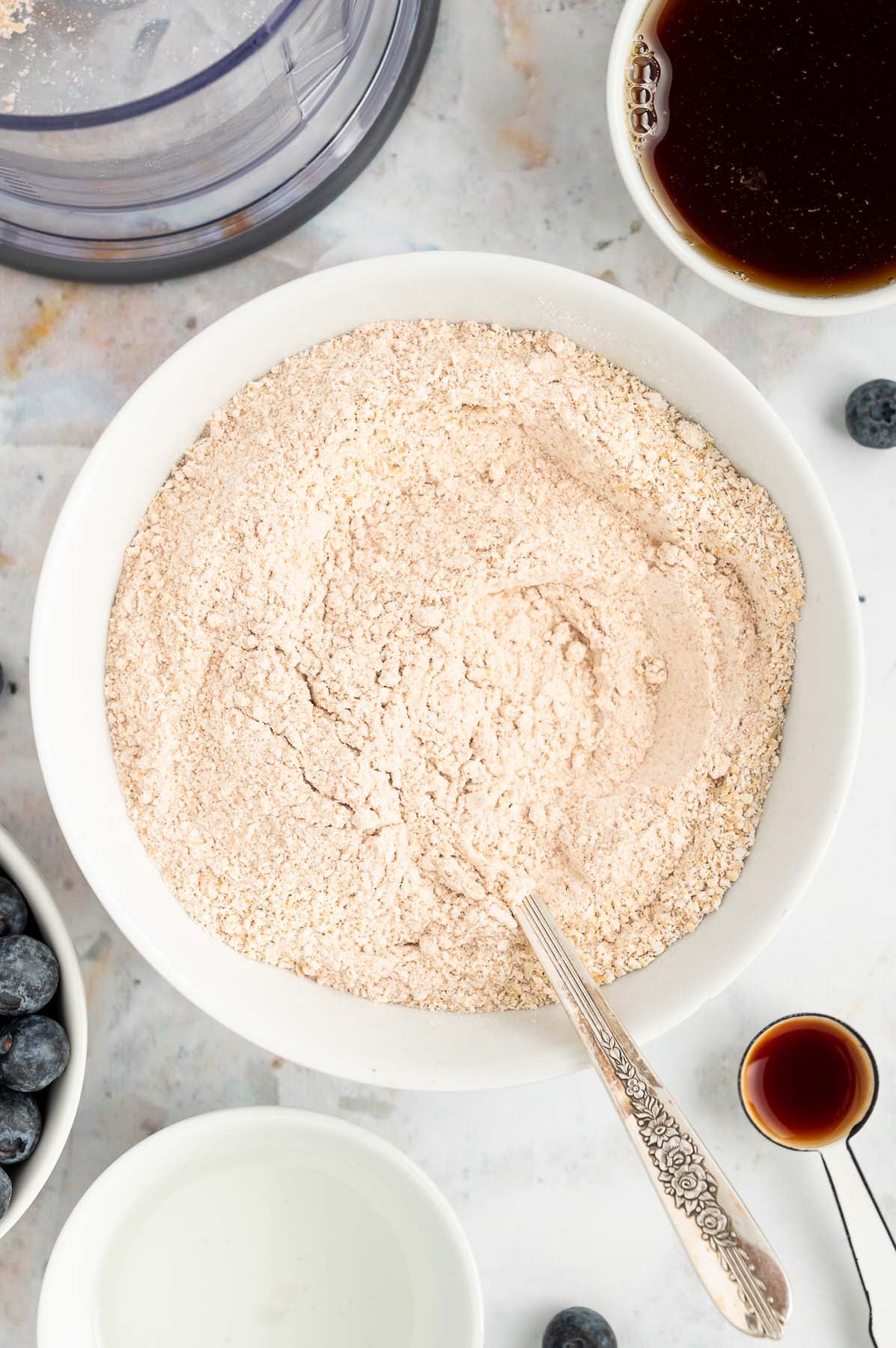An overhead of a white bowl of dry ingredients (oat flour, whole wheat flour, baking soda, baking powder, and salt) after being mixed with a silver spoon for blueberry banana muffins with more ingredients surrounding it in white bowls