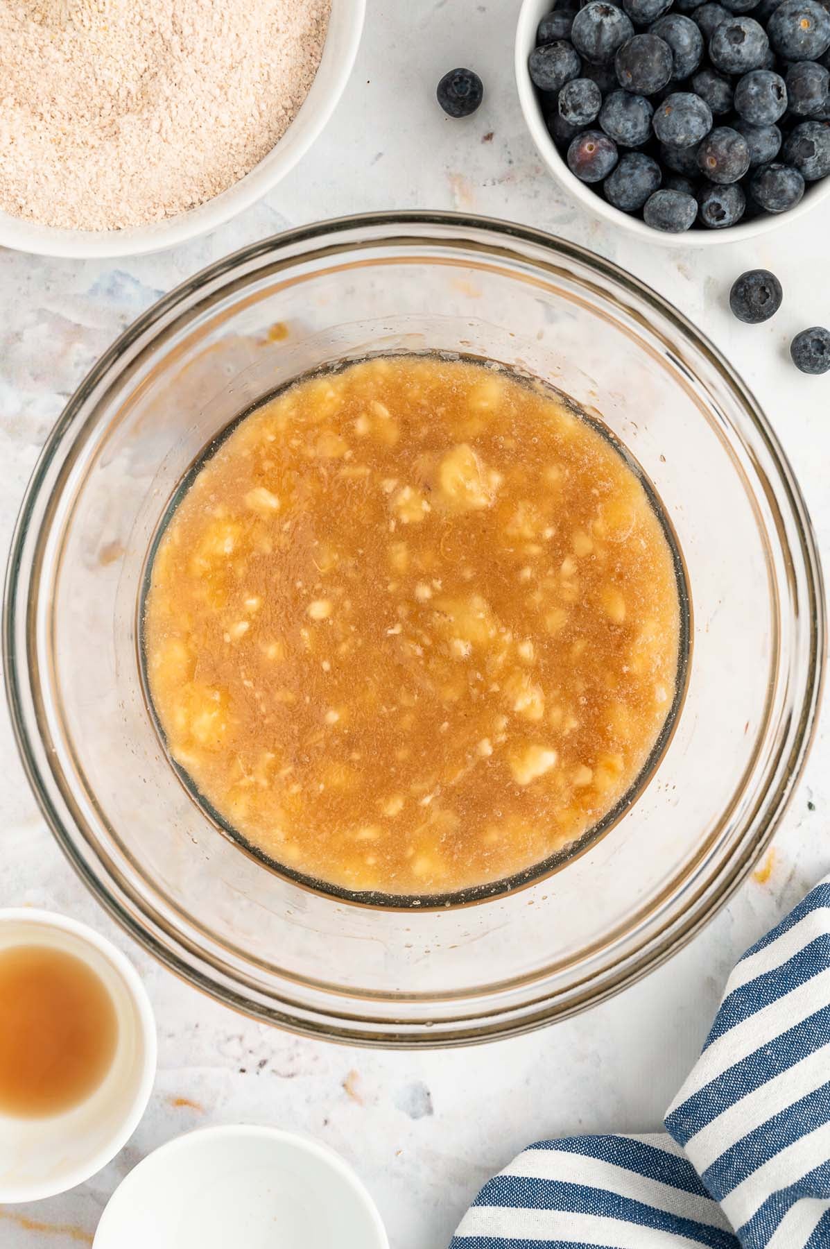 An overhead of wet ingredients (mashed bananas, eggs, maple syrup, coconut oil, and vanilla) for blueberry banana muffins in a clear bowl after mixing with dry ingredients and blueberries in bowls on the side on a white background