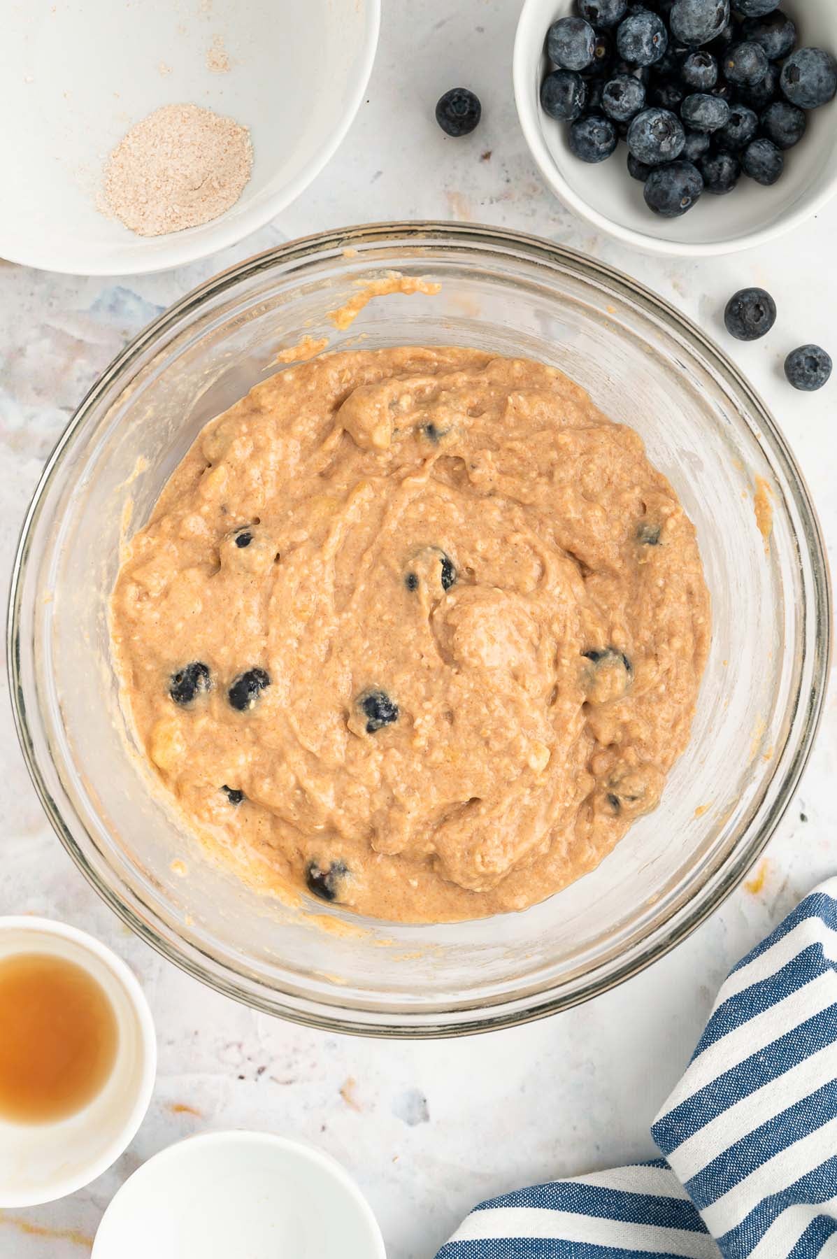 An overhead of a clear bowl with blueberry banana muffin batter in it with a white bowl of blueberries on the side on a white background