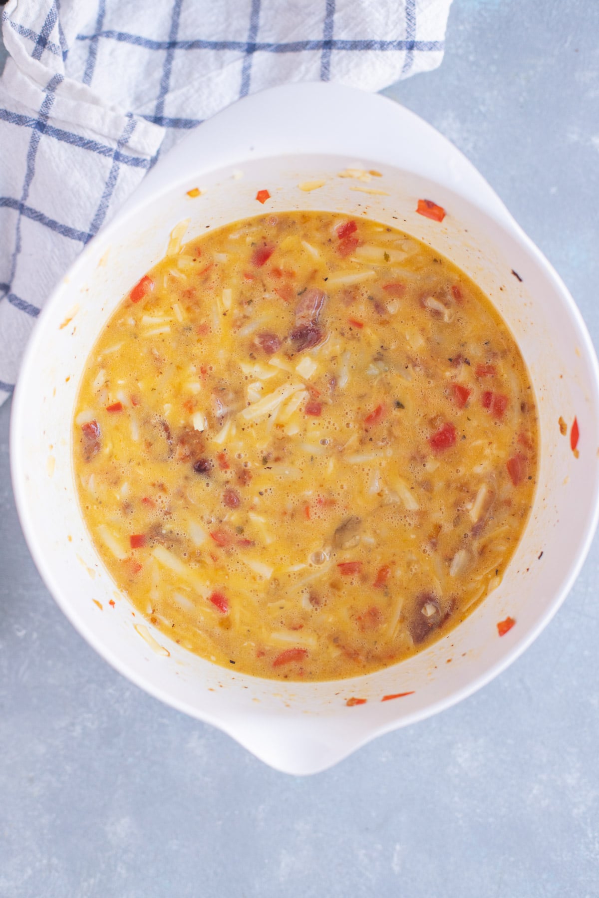 Beaten eggs in a white mixing bowl with diced red bell peppers, diced onions, and shredded cheese.