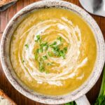 An overhead of a grey bowl of creamy vegetable soup with cream swirled in and fresh herbs on top on a wood background