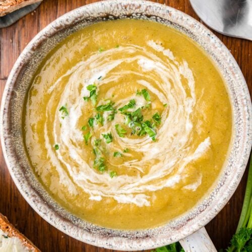 An overhead of a grey bowl of creamy vegetable soup with cream swirled in and fresh herbs on top on a wood background