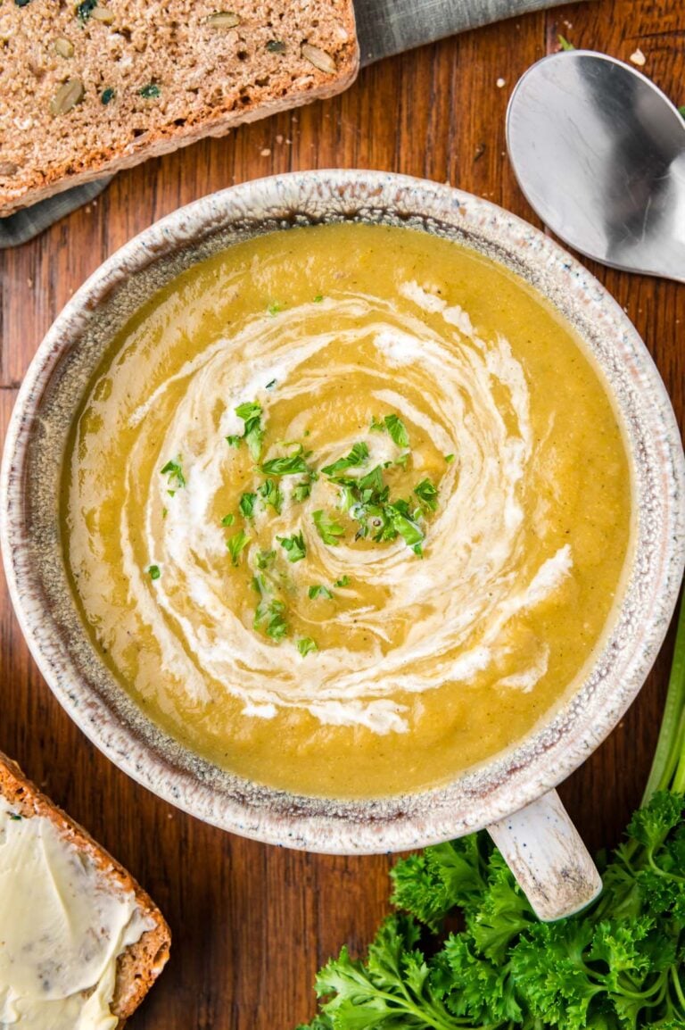An overhead of a grey bowl of creamy vegetable soup with cream swirled in and fresh herbs on top on a wood background