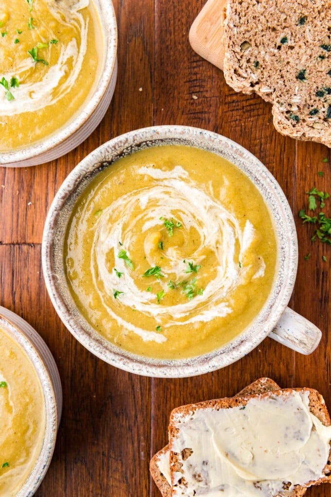 An overhead of a grey bowl of creamy vegetable soup with cream swirled in and fresh herbs on top with more bowls of soup on the side on a wood background