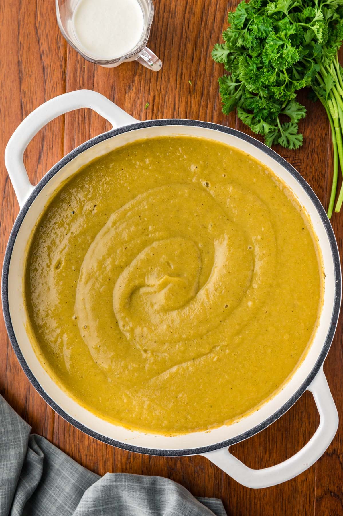 An overhead shot of a dutch oven with creamy veggie soup in it with fresh parsley on the side on a wooden background