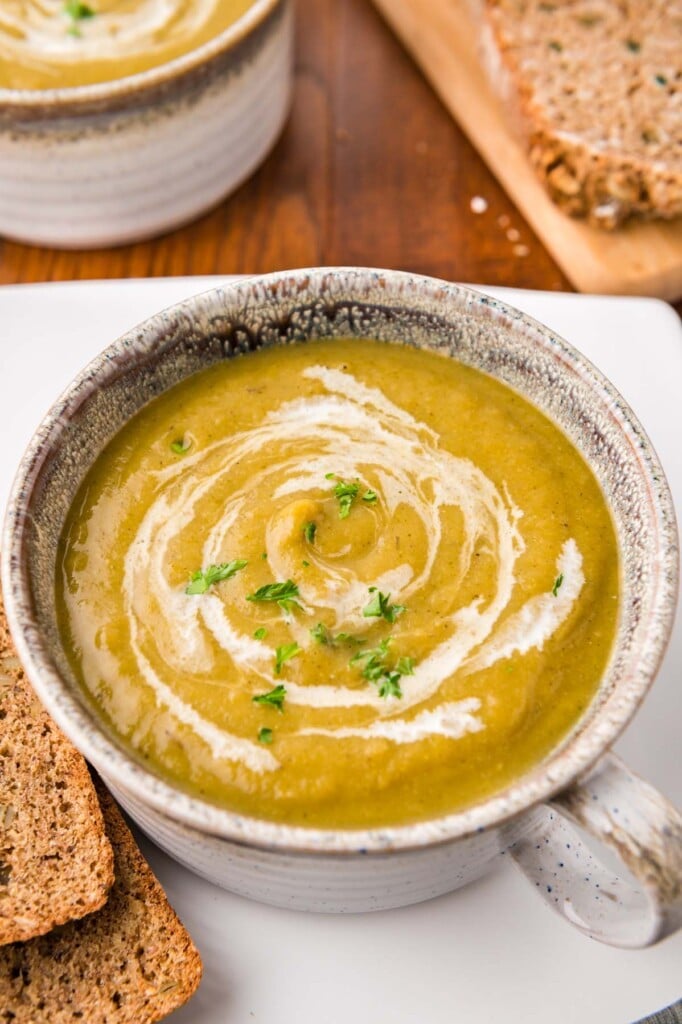 A grey bowl of creamy veggie soup with cream swirled in and fresh herbs on top on a wood background with slices of bread on the side