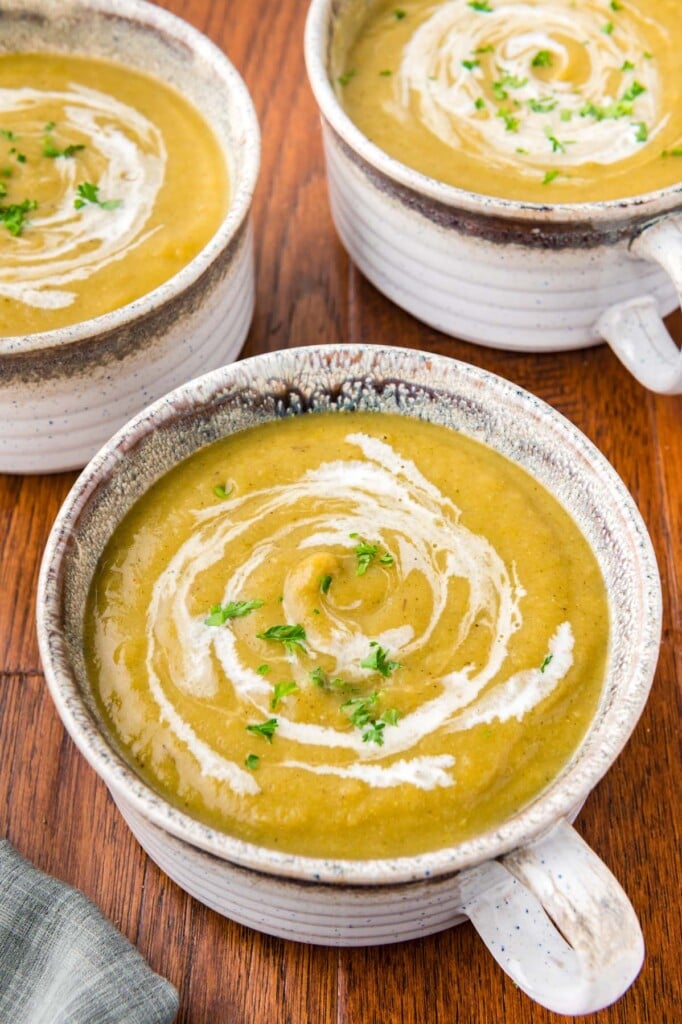 A grey bowl of creamy vegetable soup with cream swirled in and fresh herbs on top with more bowls in the background on a wood table