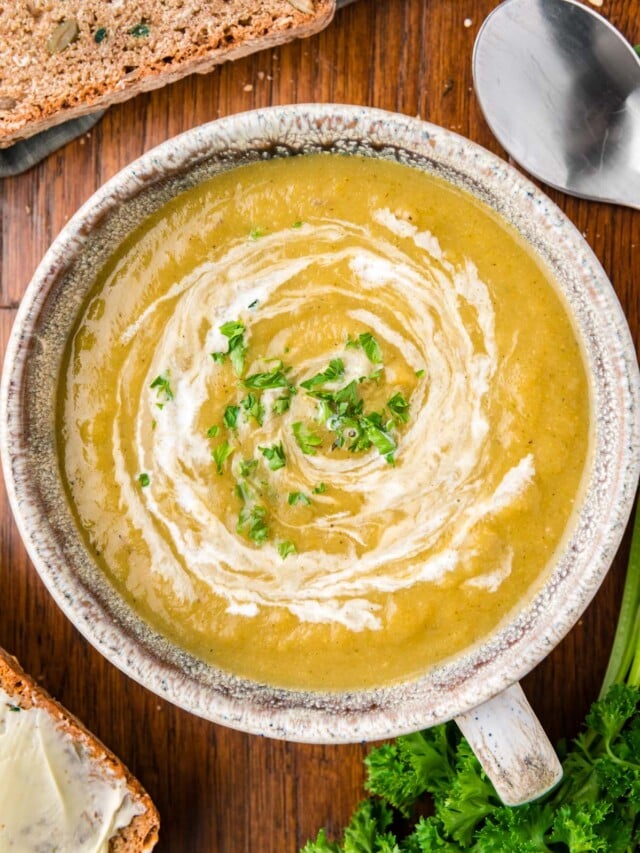 An overhead of a grey bowl of creamy vegetable soup with cream swirled in and fresh herbs on top on a wood background