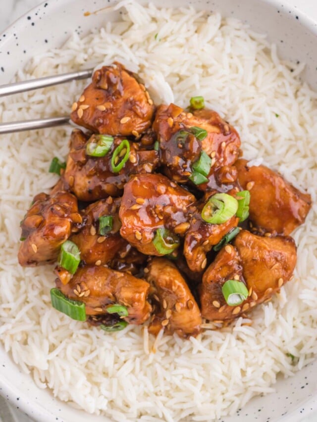 A white bowl of white rice topped with healthy ginger chicken with green onions on top and chopsticks on the side