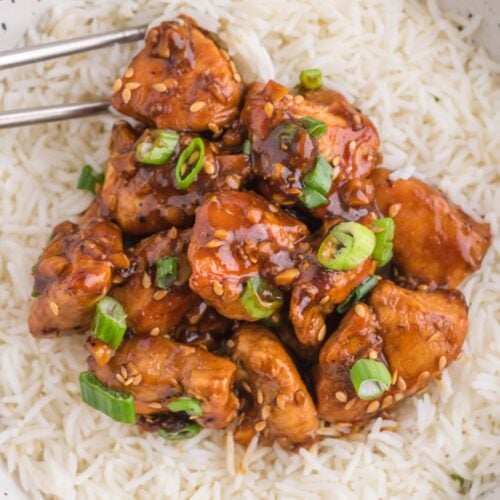 A white bowl of white rice topped with healthy ginger chicken with green onions on top and chopsticks on the side