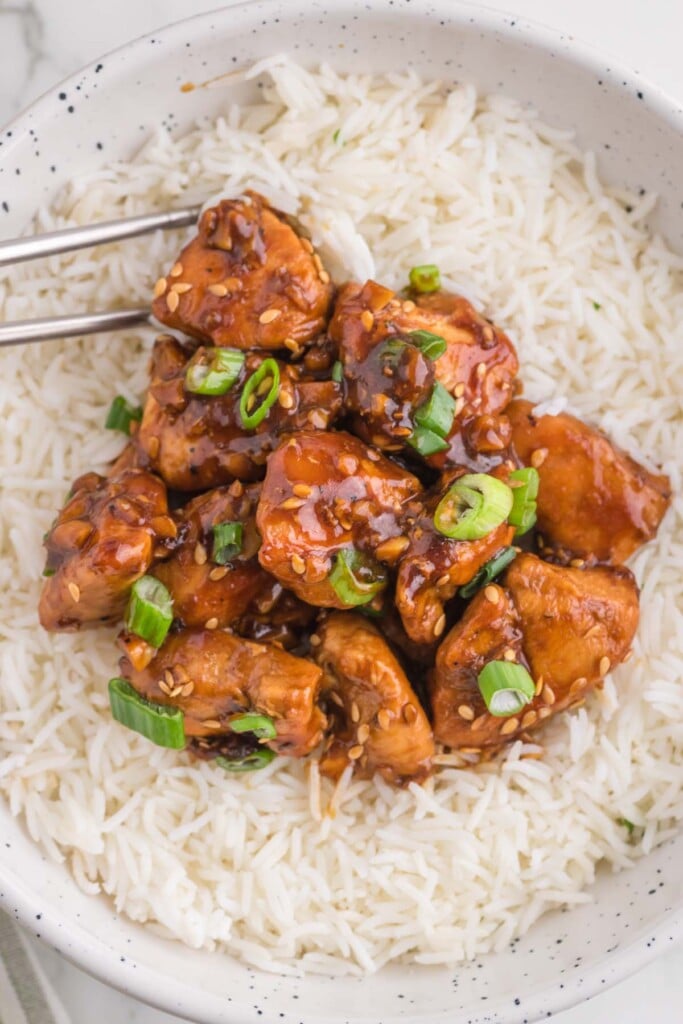 A white bowl of white rice topped with healthy ginger chicken with green onions on top and chopsticks on the side