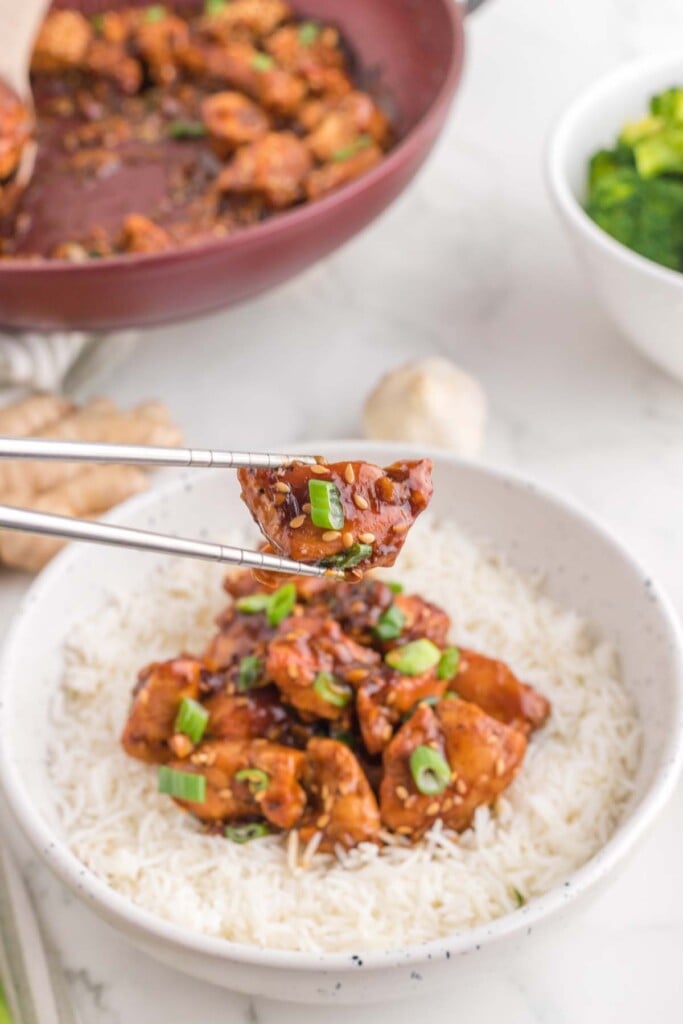 A piece of ginger chicken being picked up with chopsticks from a white bowl of ginger chicken on a bed of white rice with a pan of ginger garlic chicken in the background on a white background