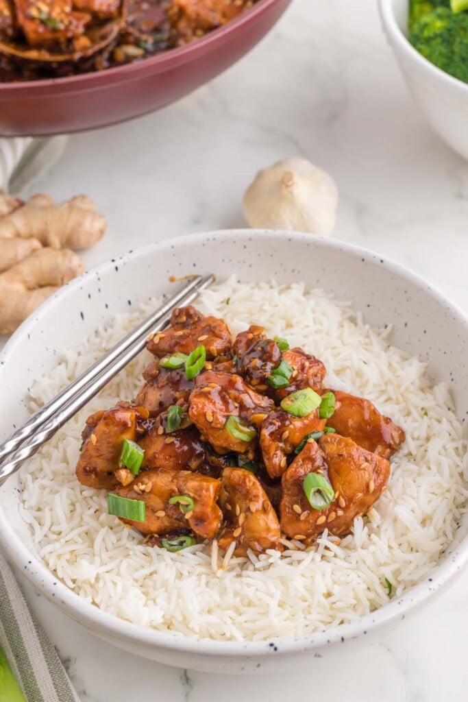 A white bowl with ginger chicken on top of white rice topped with green onions with chopsticks on the side on a white background