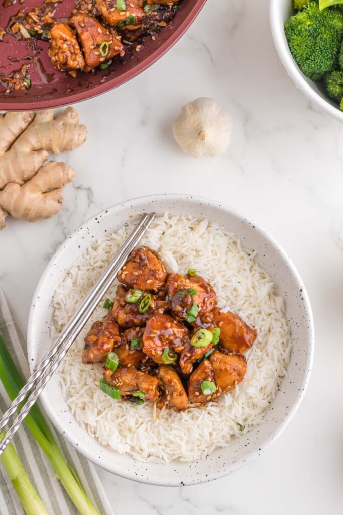 An overhead shot of a white bowl of ginger chicken on top of white rice on a white background with a pan of chicken, ginger and green onions on the side