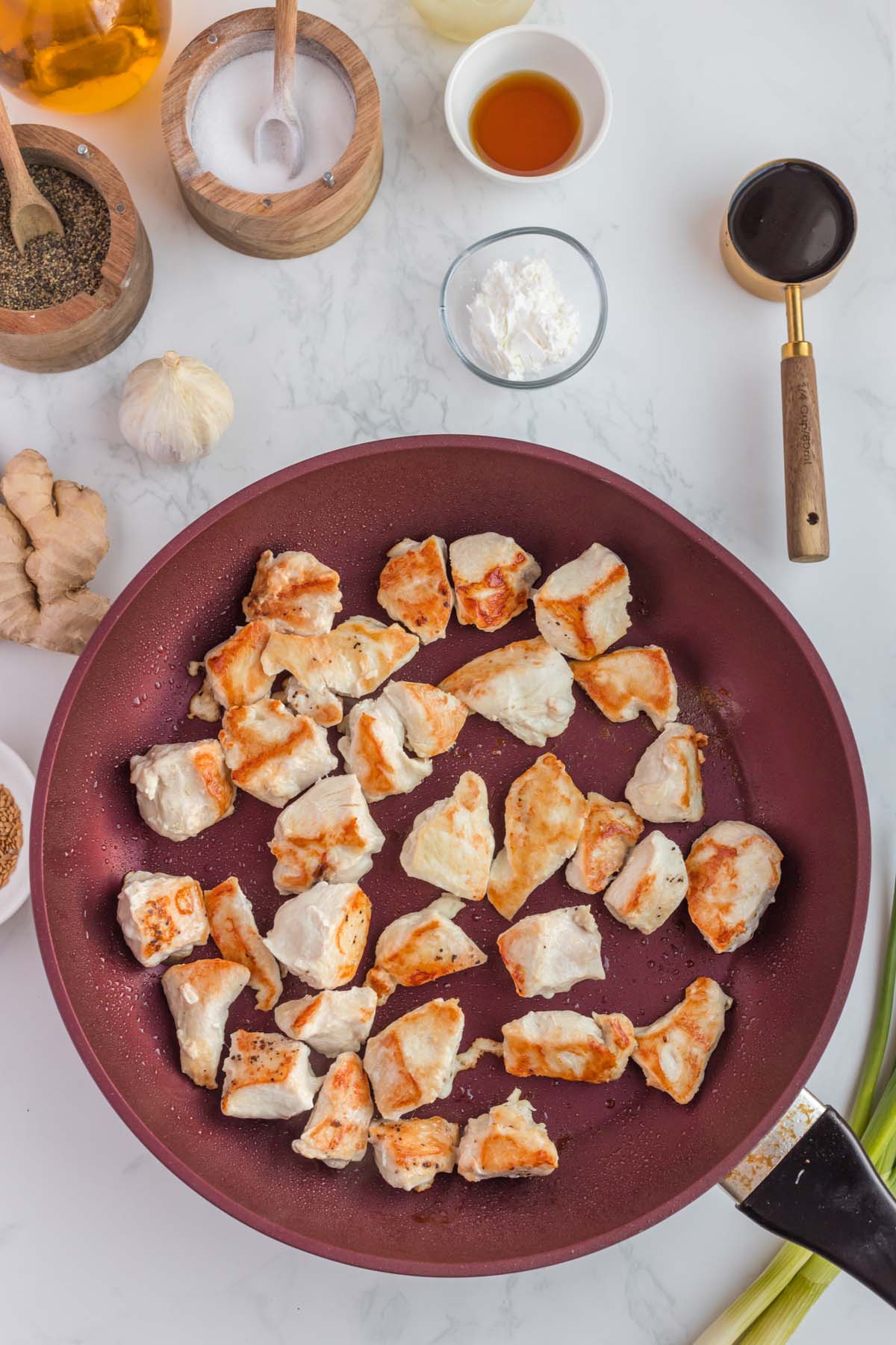 An overhead shot of a pan with browned chicken pieces for ginger chicken on a white background with more ingredients on the side