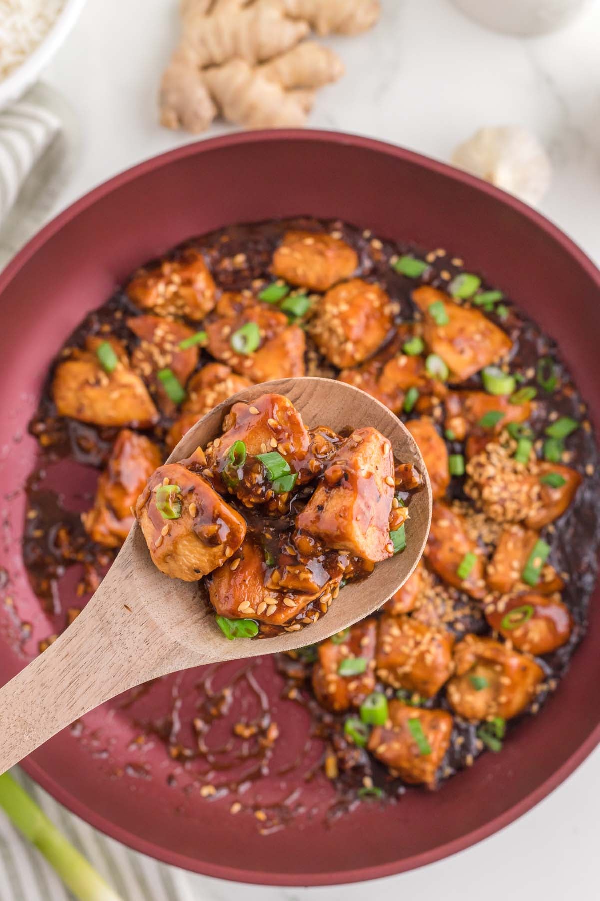 A wooden spoon taking a scoop out of a pan of ginger chicken on a white background