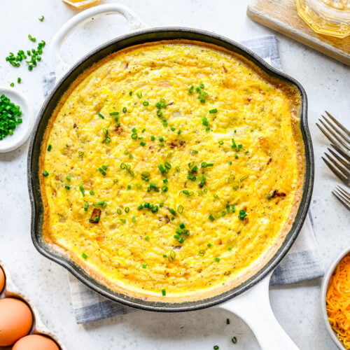An overhead shot of a cast iron pan of ham and cheese frittata topped with fresh herbs on a white background