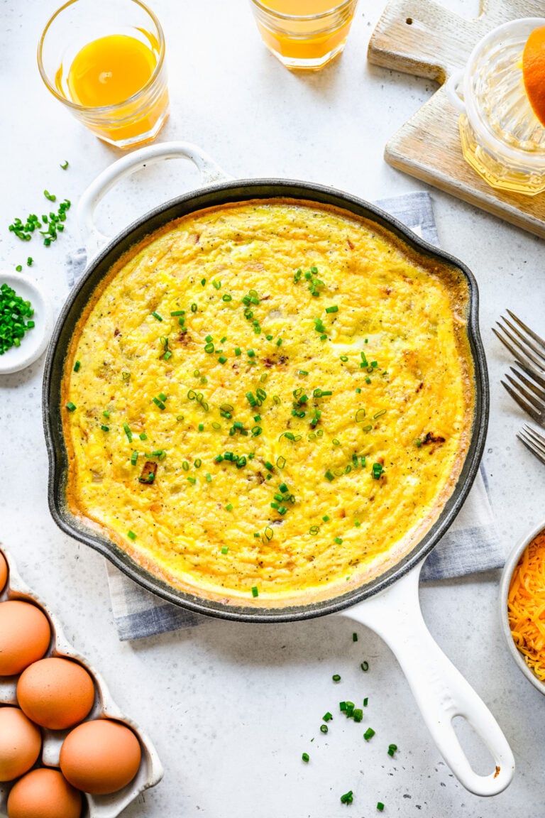 An overhead shot of a cast iron pan of ham and cheese frittata topped with fresh herbs on a white background