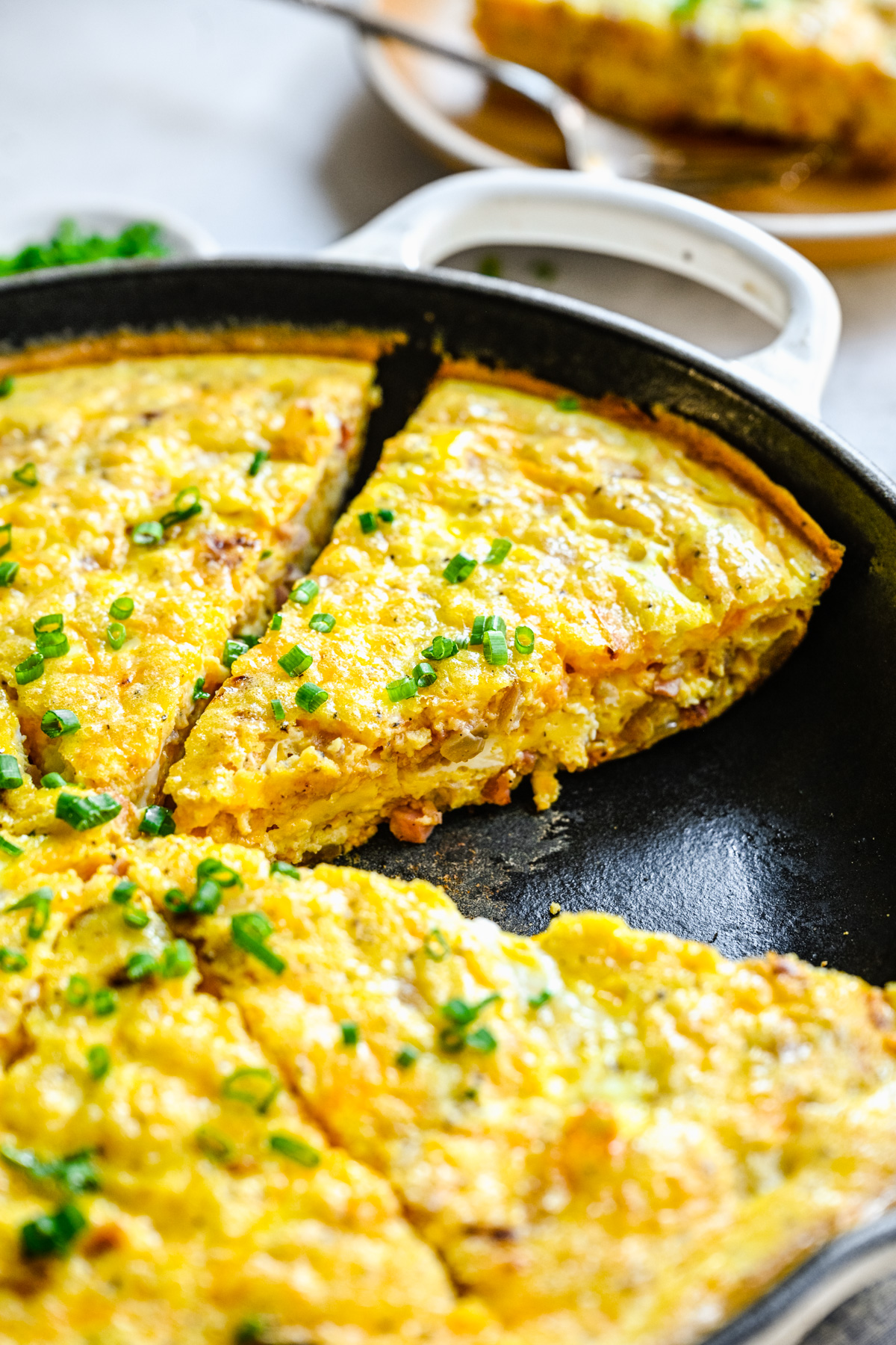 A cast iron pan of ham and cheese frittata topped with fresh chives. A slice of the baked egg dish has been removed.