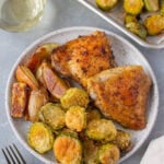 An overhead shot of a plate with parmesan brussel sprouts, roasted fingerling potatoes, and baked chicken thighs with a sheet pan of brussels, a glass of wine, and gold silverware on the side