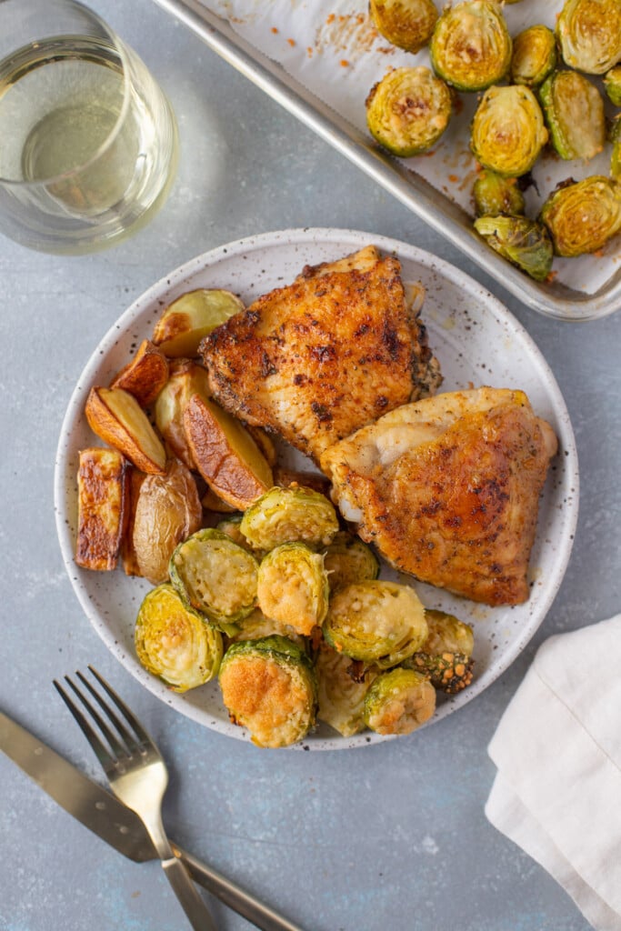 An overhead shot of a plate with parmesan brussel sprouts, roasted fingerling potatoes, and baked chicken thighs with a sheet pan of brussels, a glass of wine, and gold silverware on the side