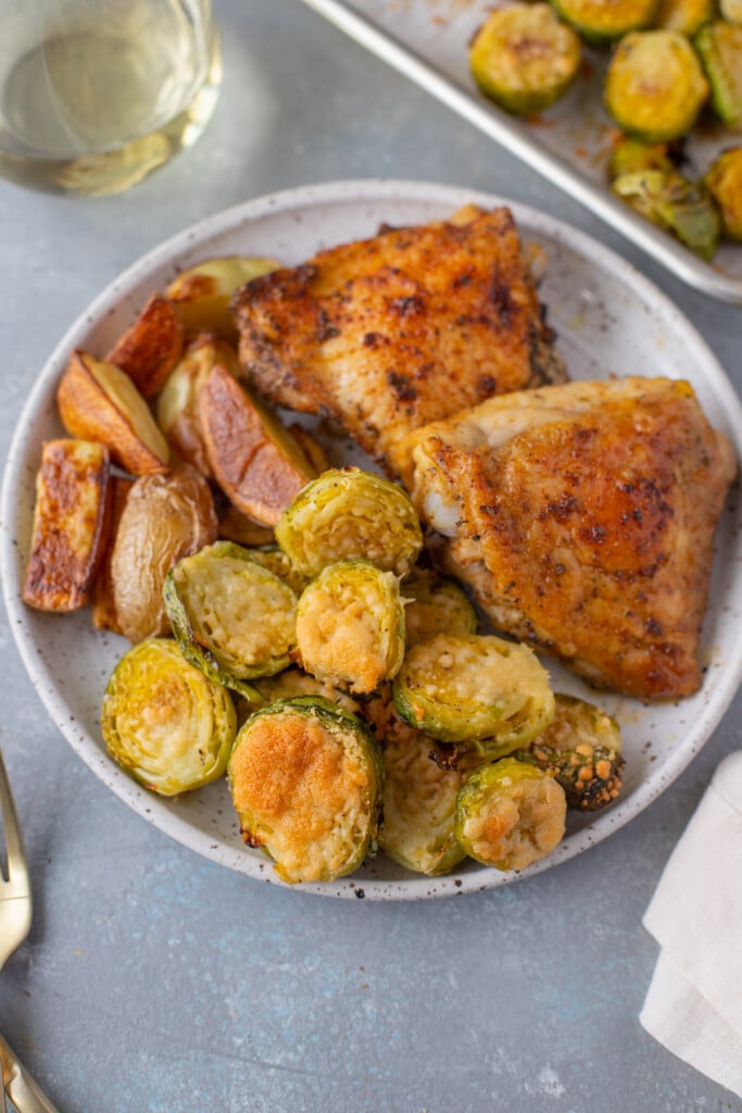 An overhead shot of a plate with parmesan brussel sprouts, roasted fingerling potatoes, and baked chicken thighs with a sheet pan of brussels and a glass of wine on the side