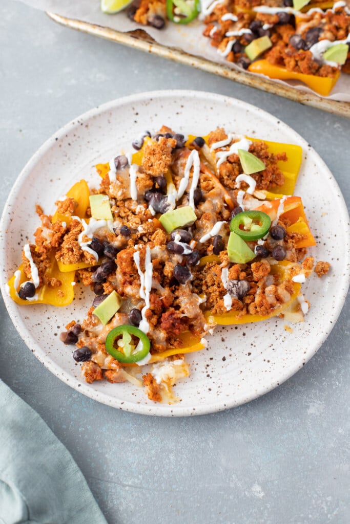 Bell pepper nachos on a white speckled plate with a sheet pan on the side on a grey background