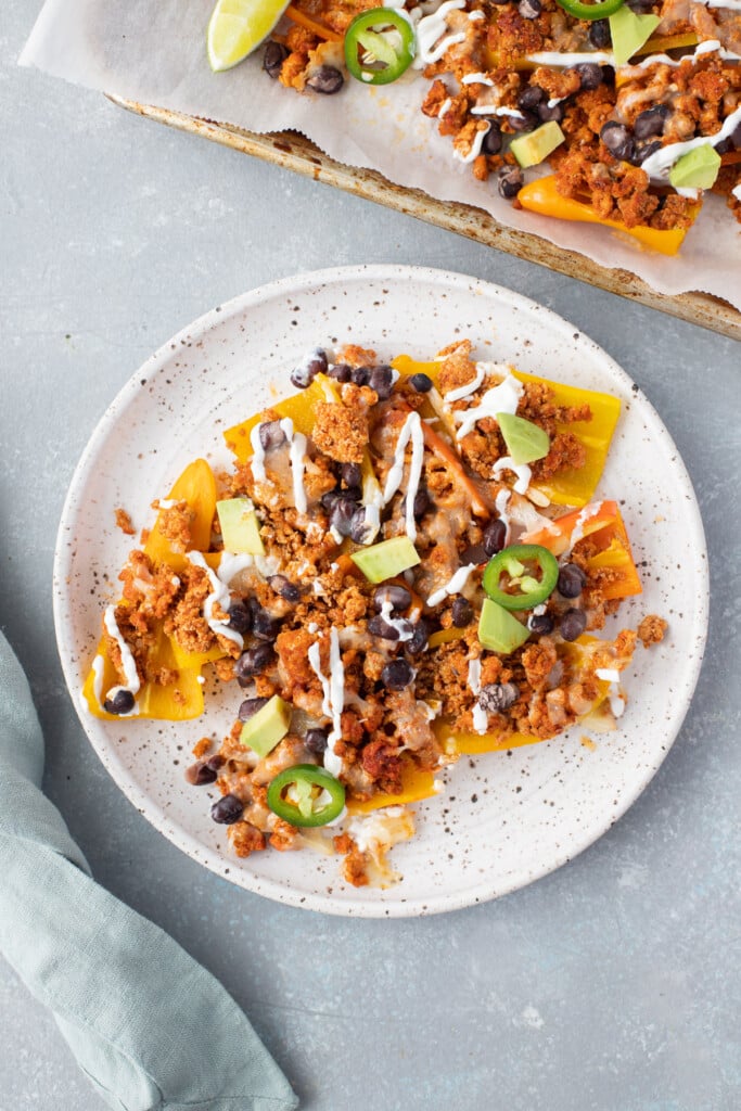 Bell pepper nachos on a white speckled plate with a sheet pan on the side on a grey background