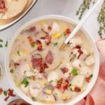 An overhead shot of chicken corn chowder in a white bowl with a spoon in it with bacon and a pot of soup on the side