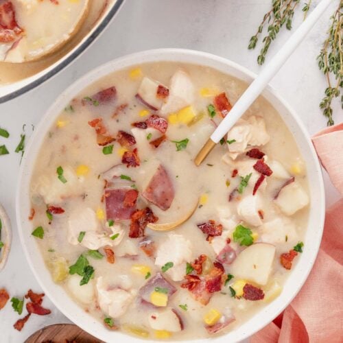 An overhead shot of chicken corn chowder in a white bowl with a spoon in it with bacon and a pot of soup on the side