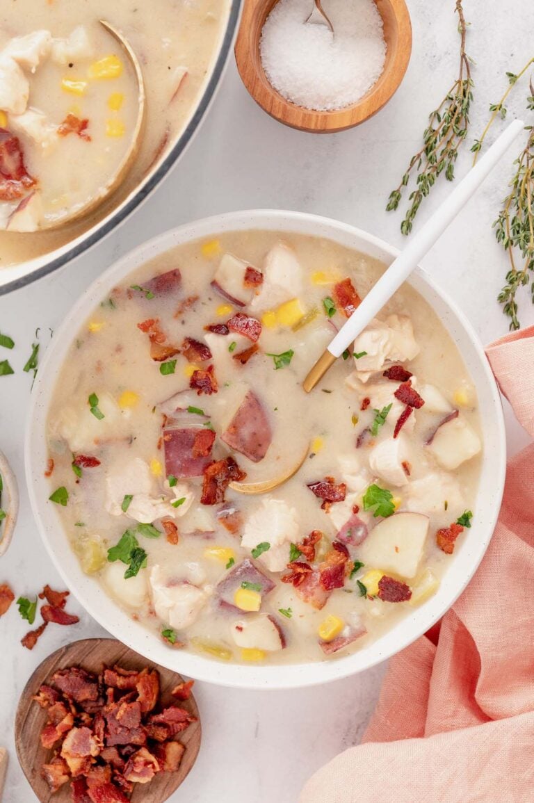 An overhead shot of chicken corn chowder in a white bowl with a spoon in it with bacon and a pot of soup on the side