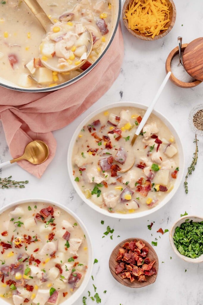 An overhead shot of two white bowls of chicken corn chowder and a dutch oven of the chowder with more ingredients on the side