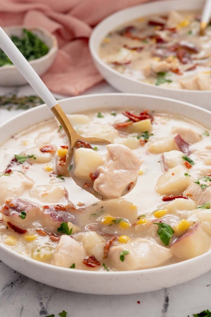 A white bowl of chicken corn chowder with a spoon taking a bite out of it with another bowl in the background