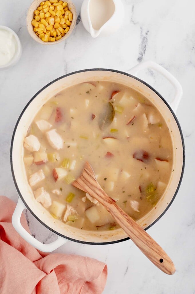 Chicken broth and chicken pieces being added to a large pot of chicken corn chowder cooking with a wooden spoon in it