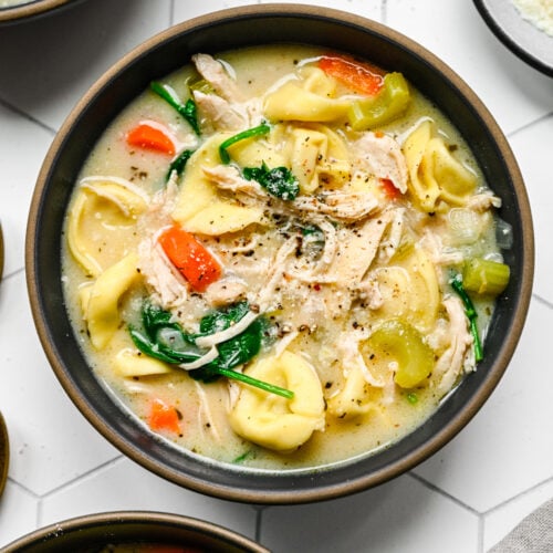 An overhead shot of a bowl of chicken tortellini soup with more bowls on the side on a white background