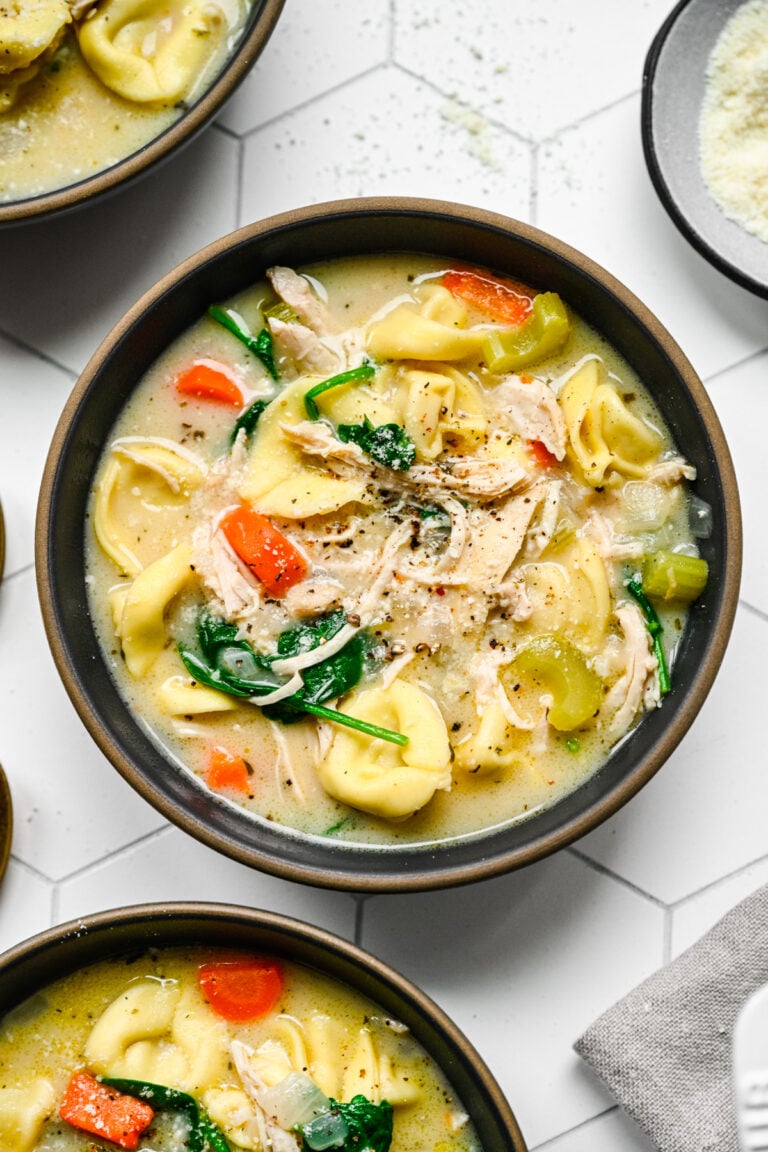An overhead shot of a bowl of chicken tortellini soup with more bowls on the side on a white background