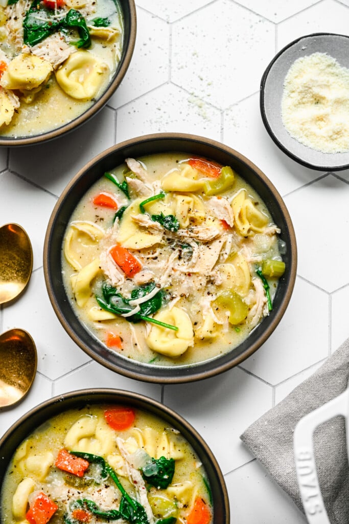 An overhead shot of a bowl of chicken tortellini soup with more bowls and spoons on the side on a white background