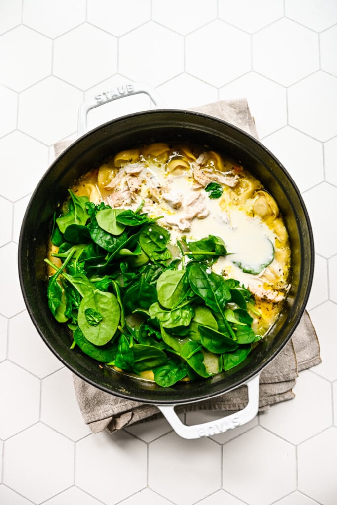 Spinach and cream being added to a pot of chicken tortellini soup on a white background