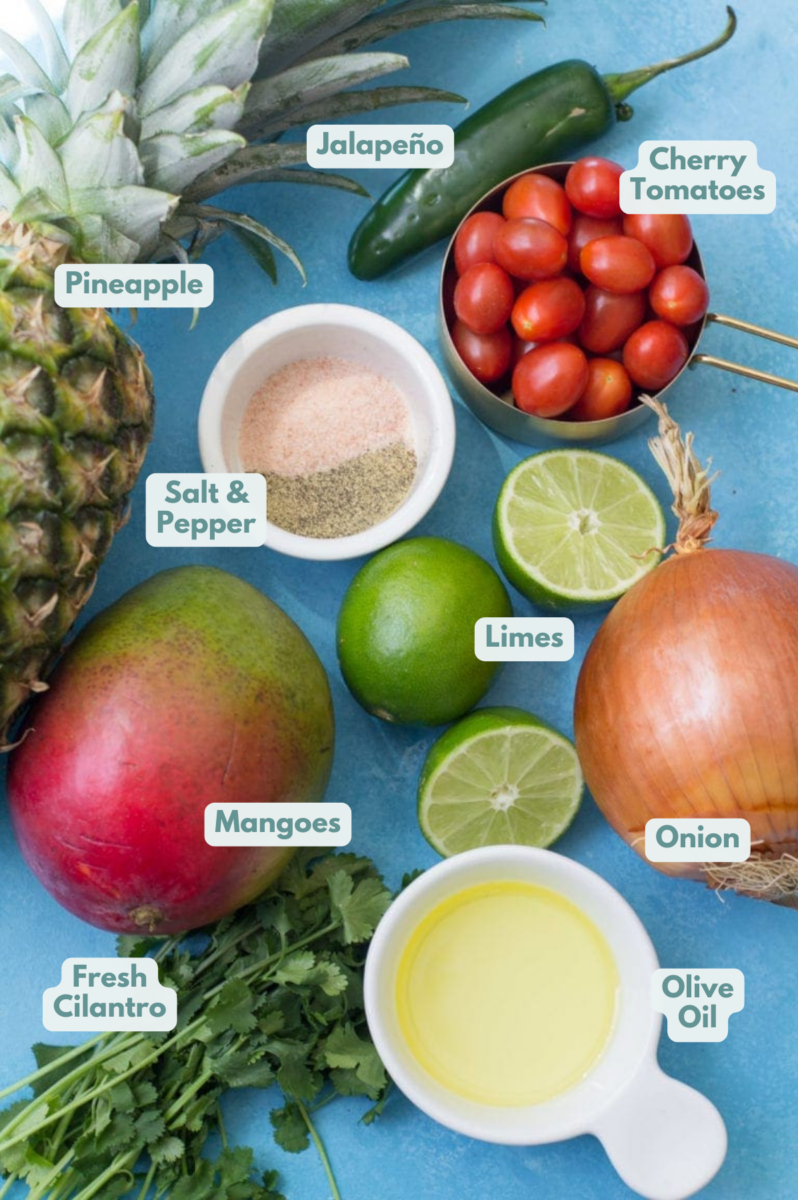 Ingredients on a blue counter, viewed from overhead: Whole pineapple, fresh mango, small bowl of salt and black pepper, 2 limes, bunch of fresh cilantro, olive oil, unpeeled yellow onion, cherry tomatoes, and a whole jalapeno.