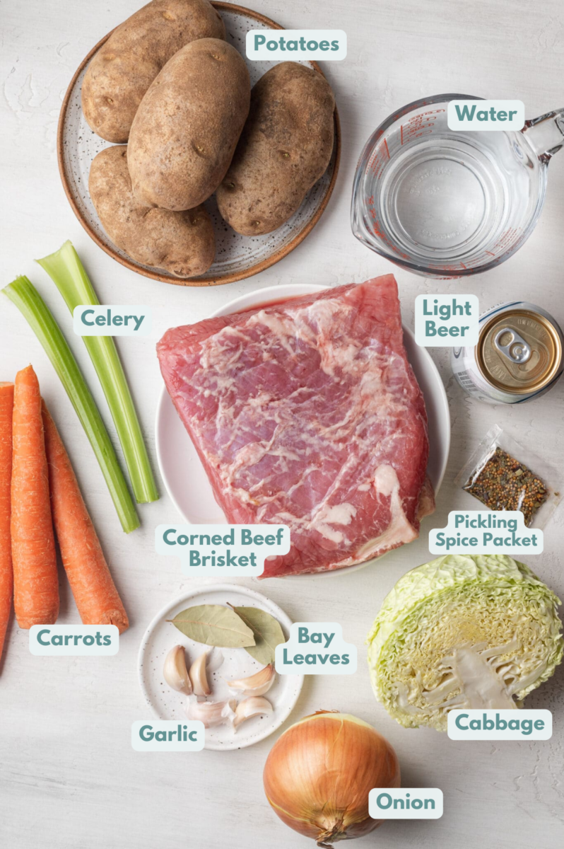 Ingredients for slow cooker corned beef in white bowls: corned beef brisket, carrots, potatoes, celery, cabbage, onion, garlic, bay leaves, light beer, water, pickling spice packet